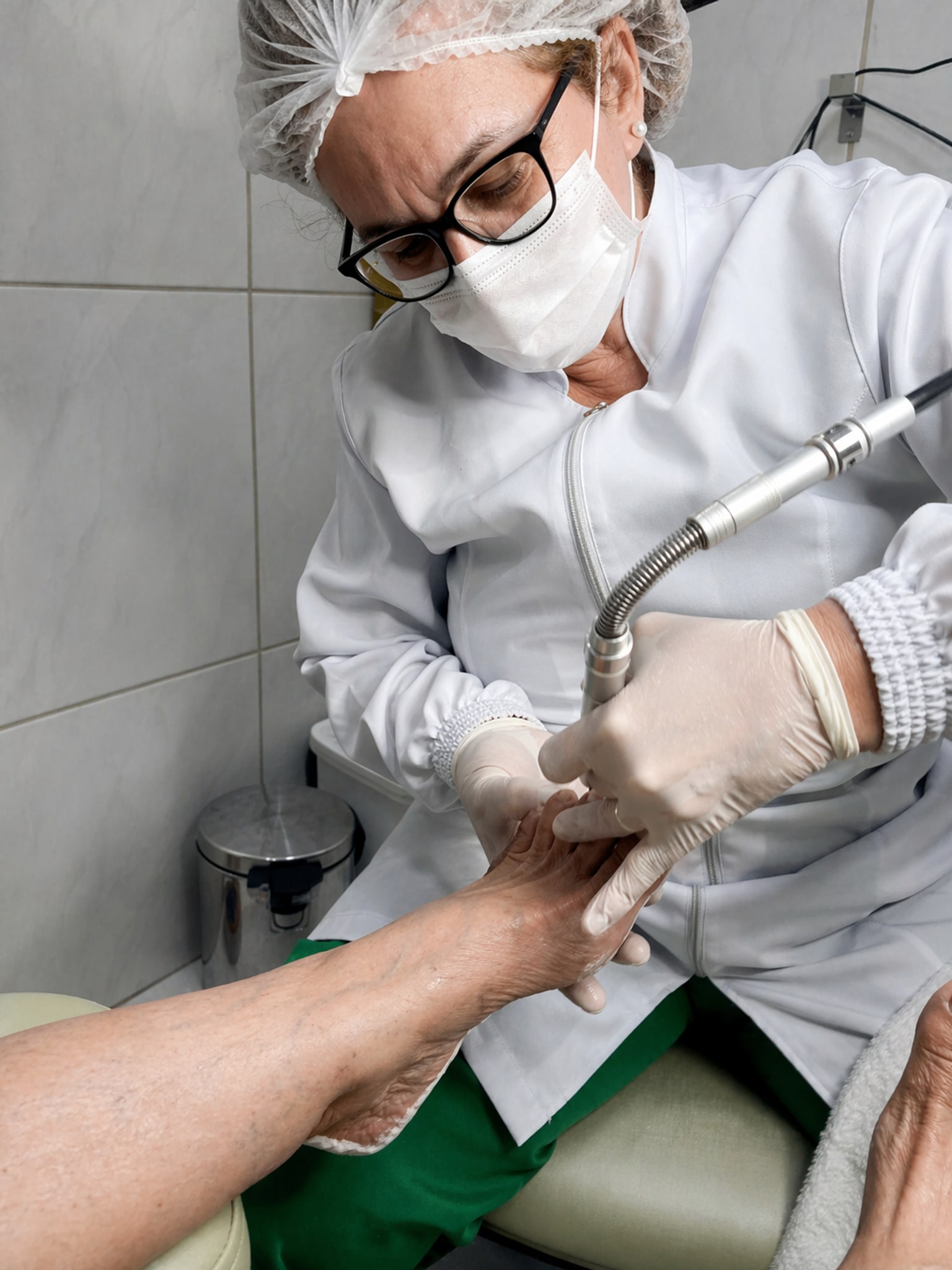 Zucarina, podóloga da ZL Podologia Fortaleza, em atendimento clínico com jaleco branco, touca, máscara, óculos e luvas, segurando o pé da paciente com precisão