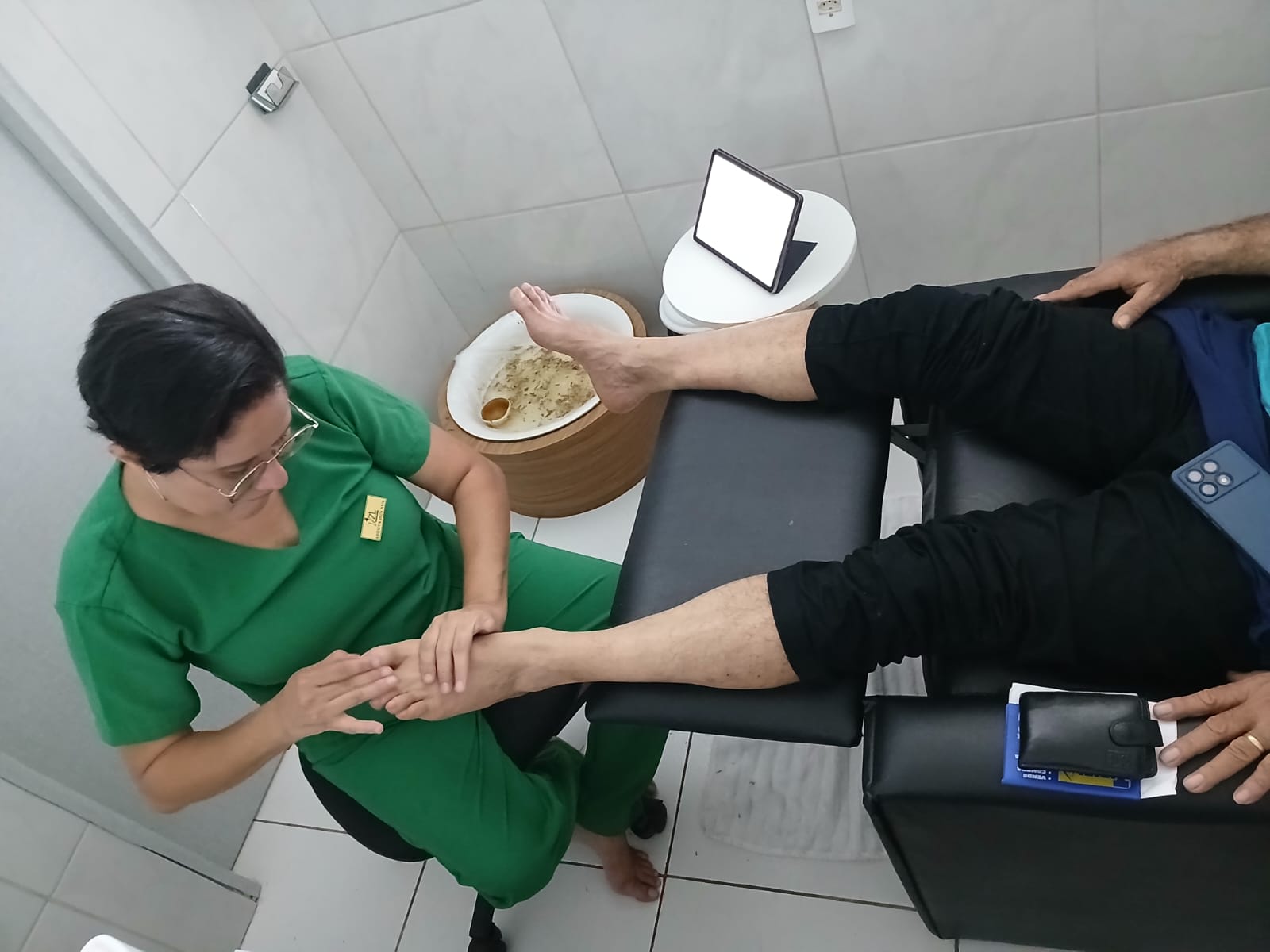 Jannie, podóloga da ZL Podologia, atendendo uma paciente na sala da clínica em Fortaleza — jaleco verde, óculos, atenção individualizada